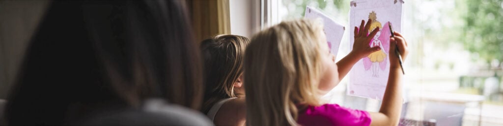 Barn ritar och sätter upp teckningar på ett fönster med dagsljus utanför.