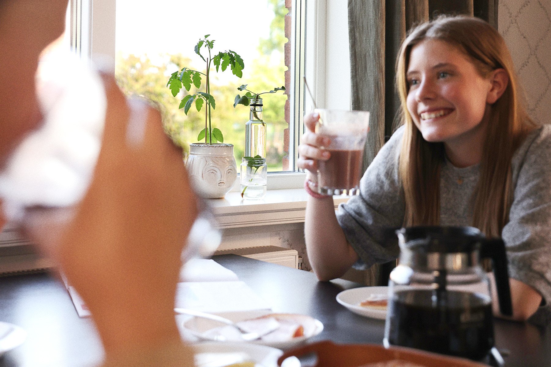 Person sitter vid fönster och dricker, med växter på fönsterbrädan.