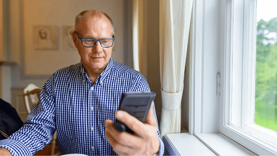 Äldre man sitter vid ett fönster och tittar på sin mobiltelefon.