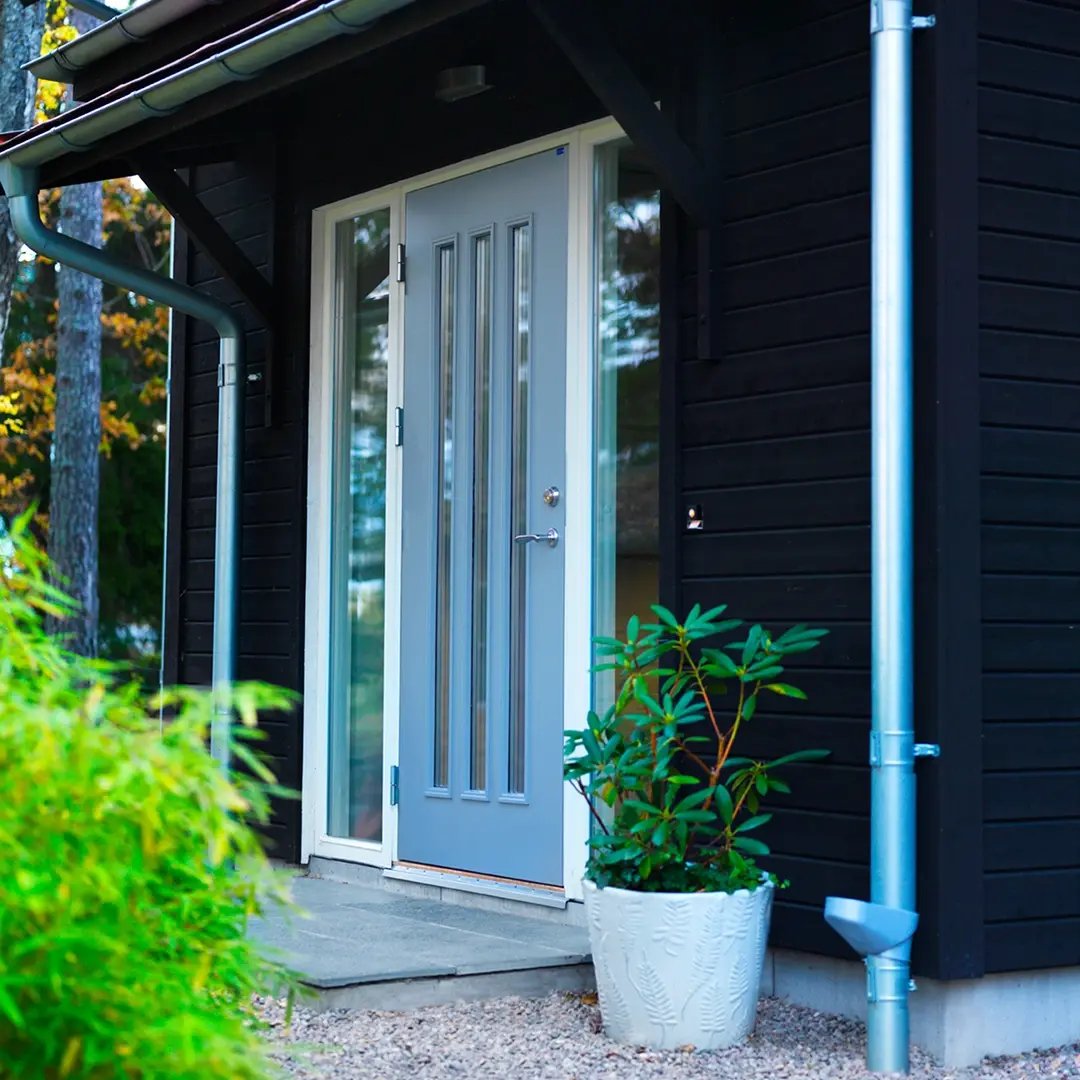 Grå ytterdörr med glaspartier på en mörk träfasad, med växt i kruka bredvid entrén.