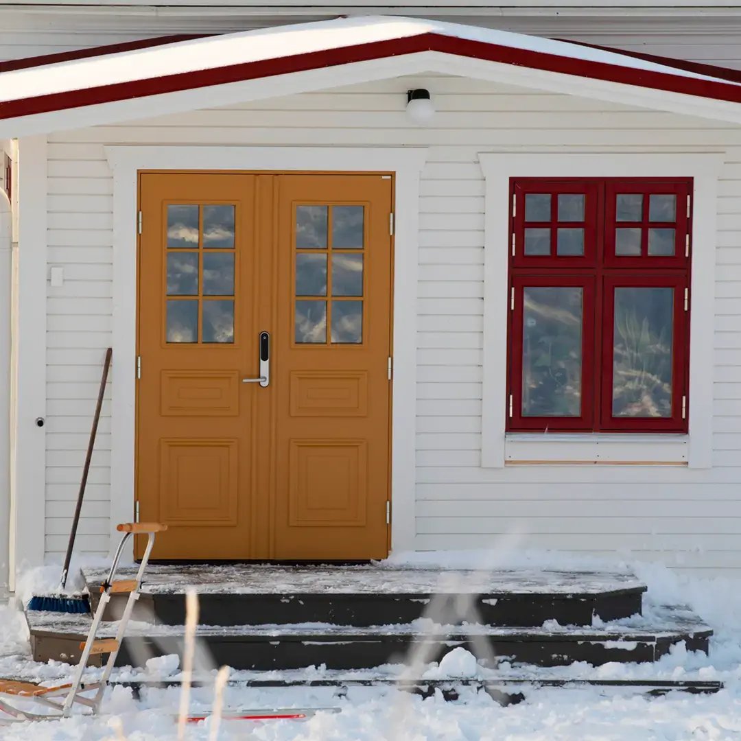 Gul ytterdörr och rött fönster på en vit träfasad med snö på trappan utanför.