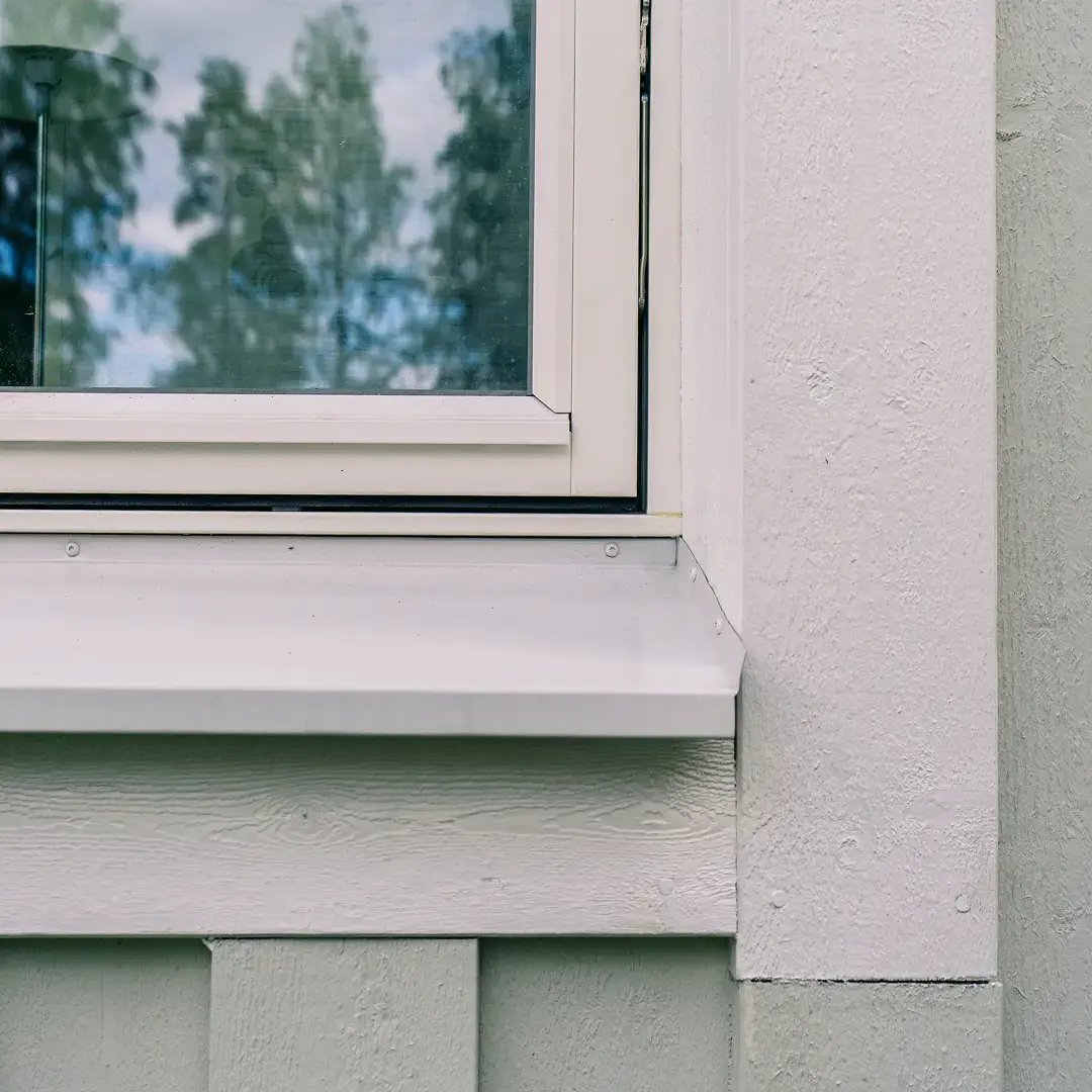 Detalj av ett vitt fönster och fönsterbleck på en ljus fasad med reflektion av träd i glaset.