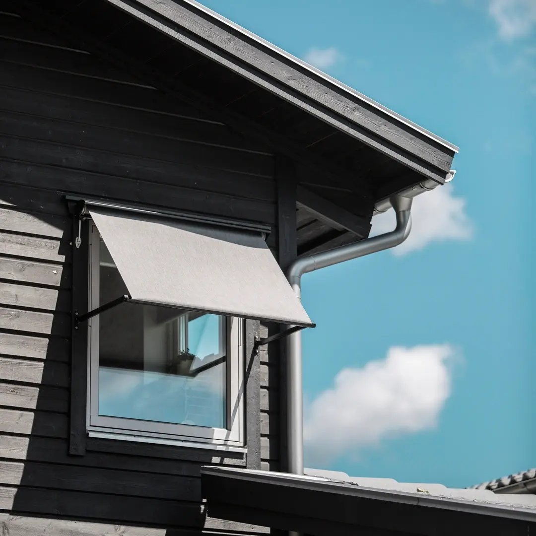 Fönster med nedfälld markis på en mörk träfasad mot blå himmel med moln.