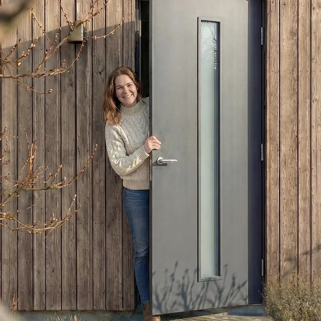 Leende kvinna öppnar en ytterdörr i en träfasad och tittar ut.