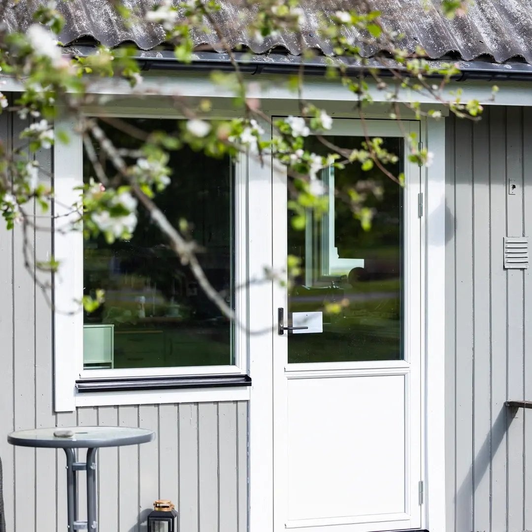 En liten grå byggnad sedd utifrån, med en vit dörr och ett fönster, delvis inramad av blommande trädgrenar i förgrunden.