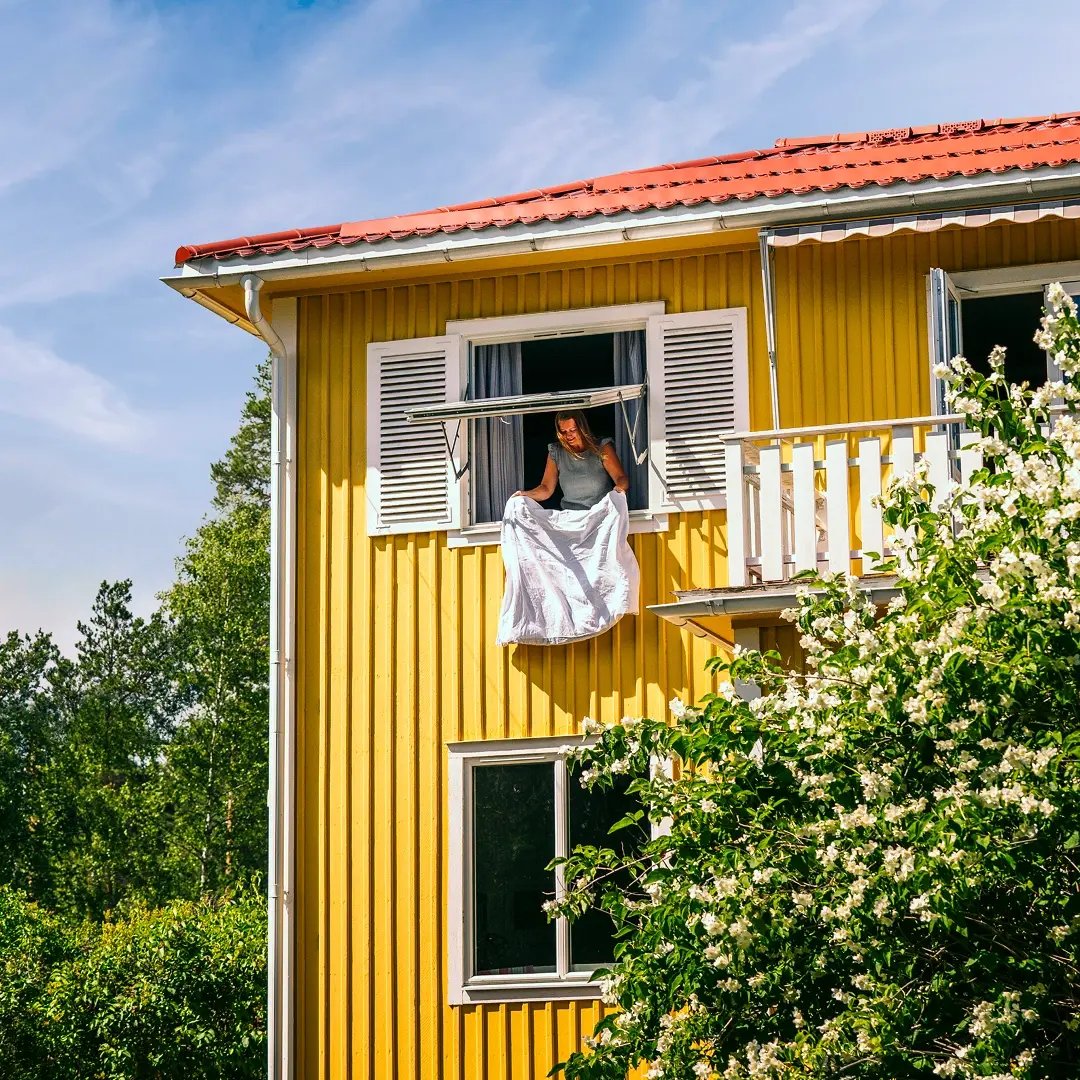 Kvinna lutar sig ut genom ett öppet fönster på övervåningen i ett gult trähus med rött tegeltak, medan hon skakar ett vitt lakan.