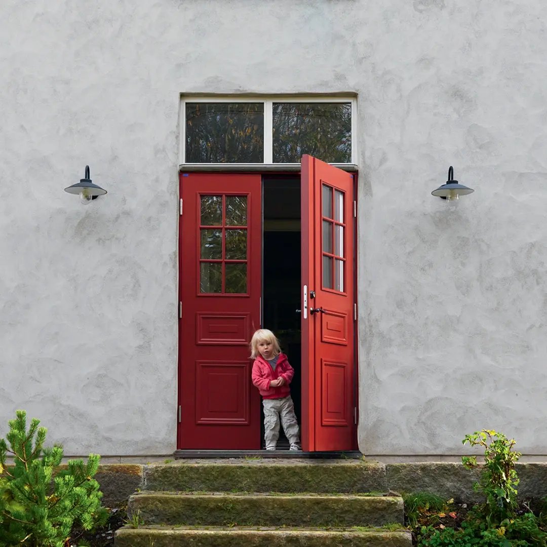 Litet barn står i en öppen röd ytterdörr till ett hus med ljus putsad fasad och trappsteg framför ingången.