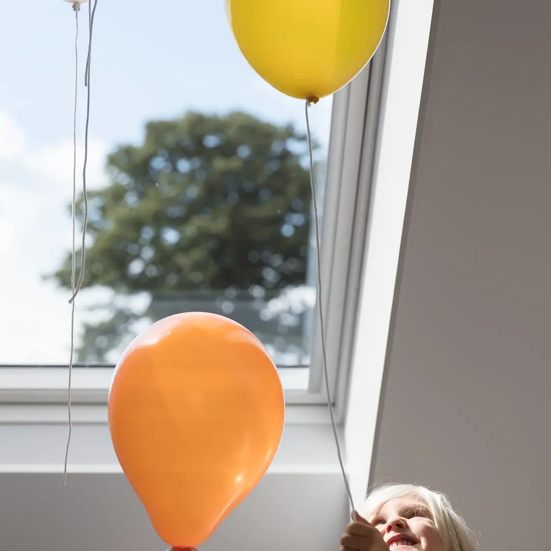 Färgglada ballonger svävar vid ett takfönster medan ett litet barn tittar upp underifrån.