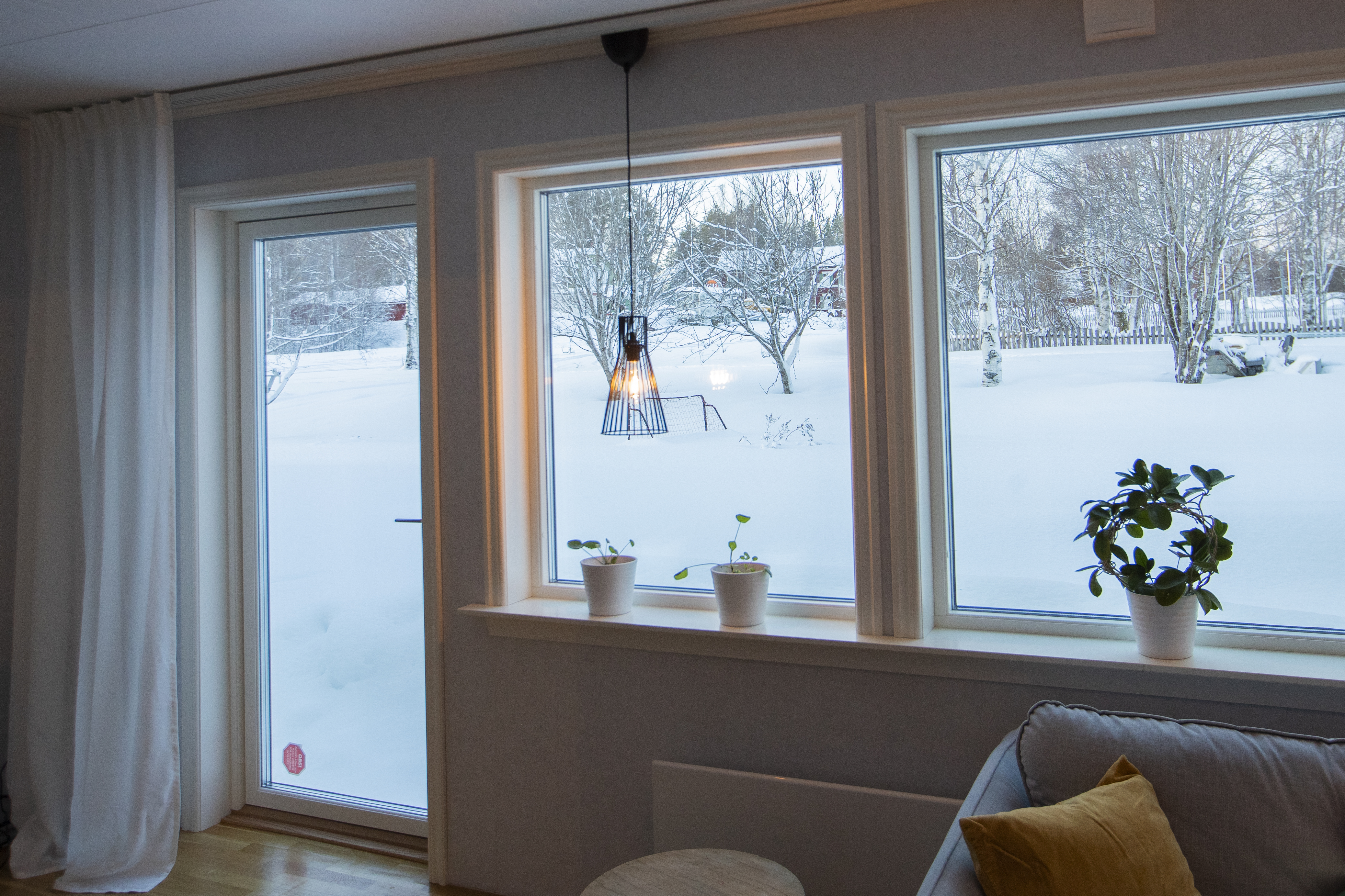 Vardagsrum med stora fönster och utsikt över snötäckt trädgård, med växter i fönsterkarmen och en hängande lampa.