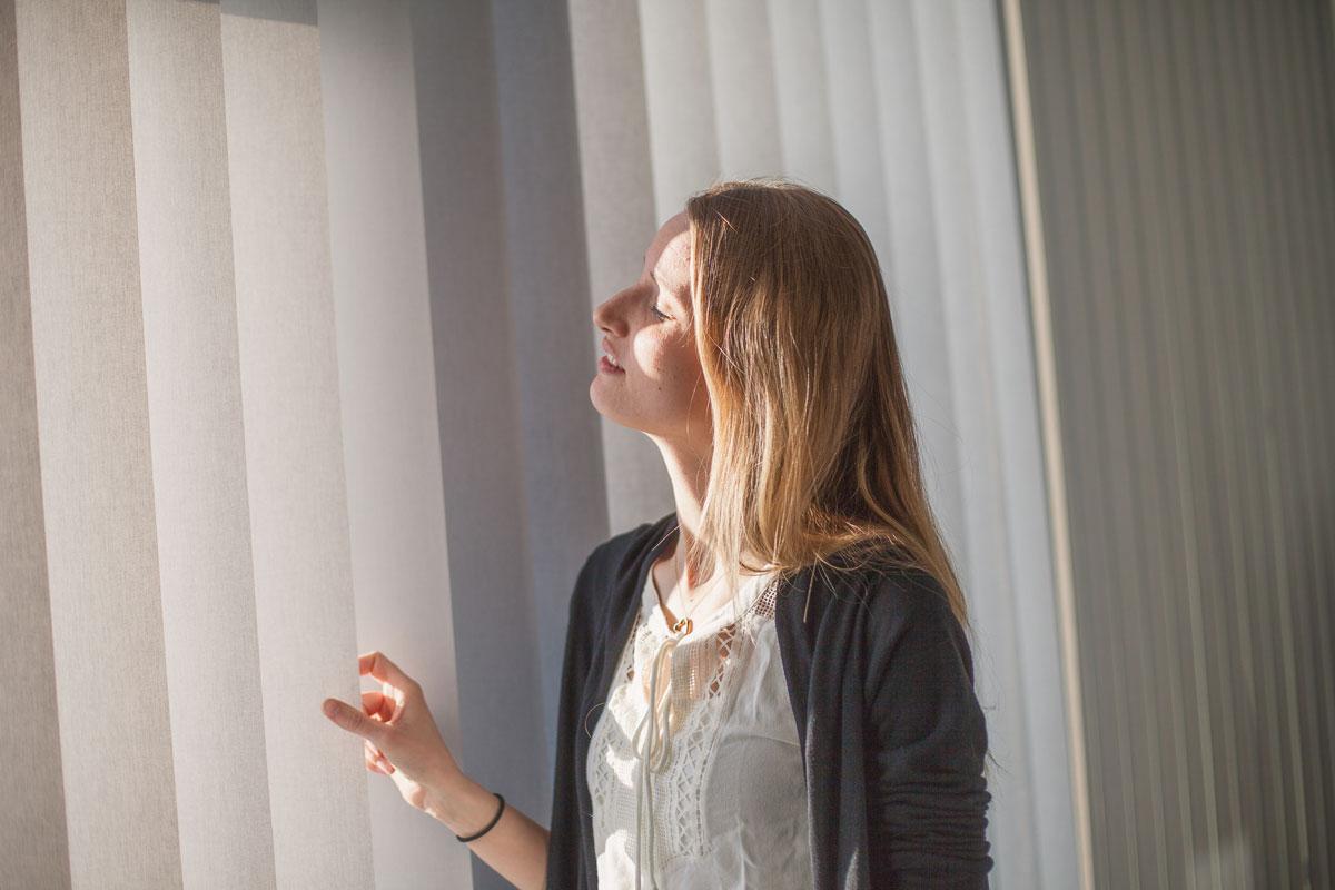 Kvinna står vid fönster och justerar gardin i dagsljus.