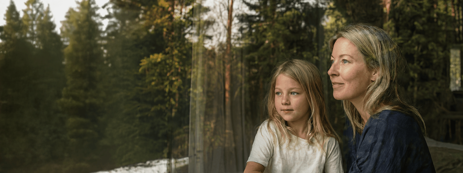 En kvinna och en ung flicka tittar ut genom ett stort fönster mot ett skogslandskap.