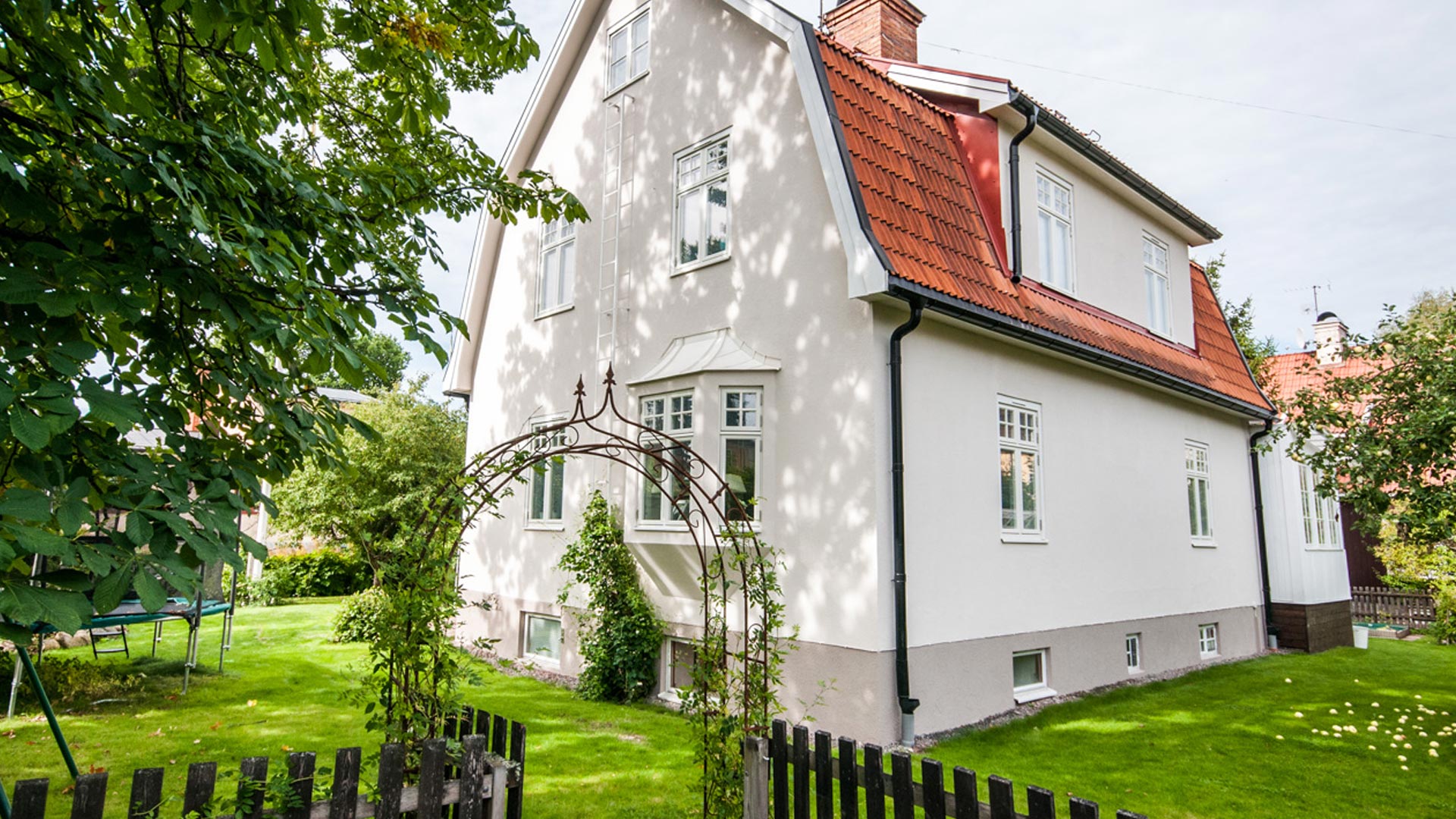 Vit villa med rött tak omgiven av grön trädgård och träd.