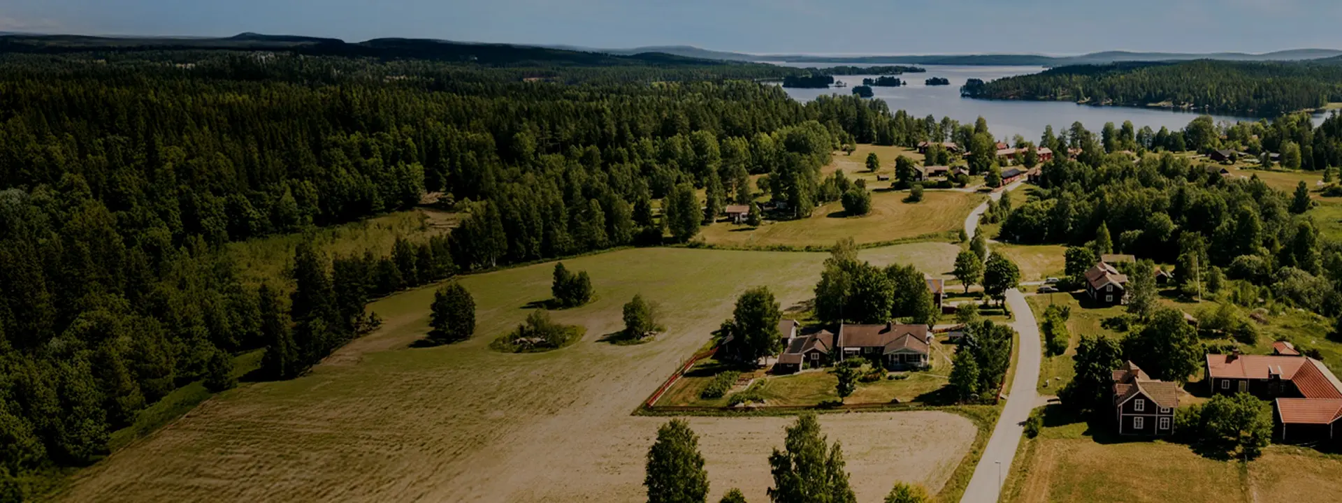 Flygbild över landskap med skog, fält och hus vid sjö.
