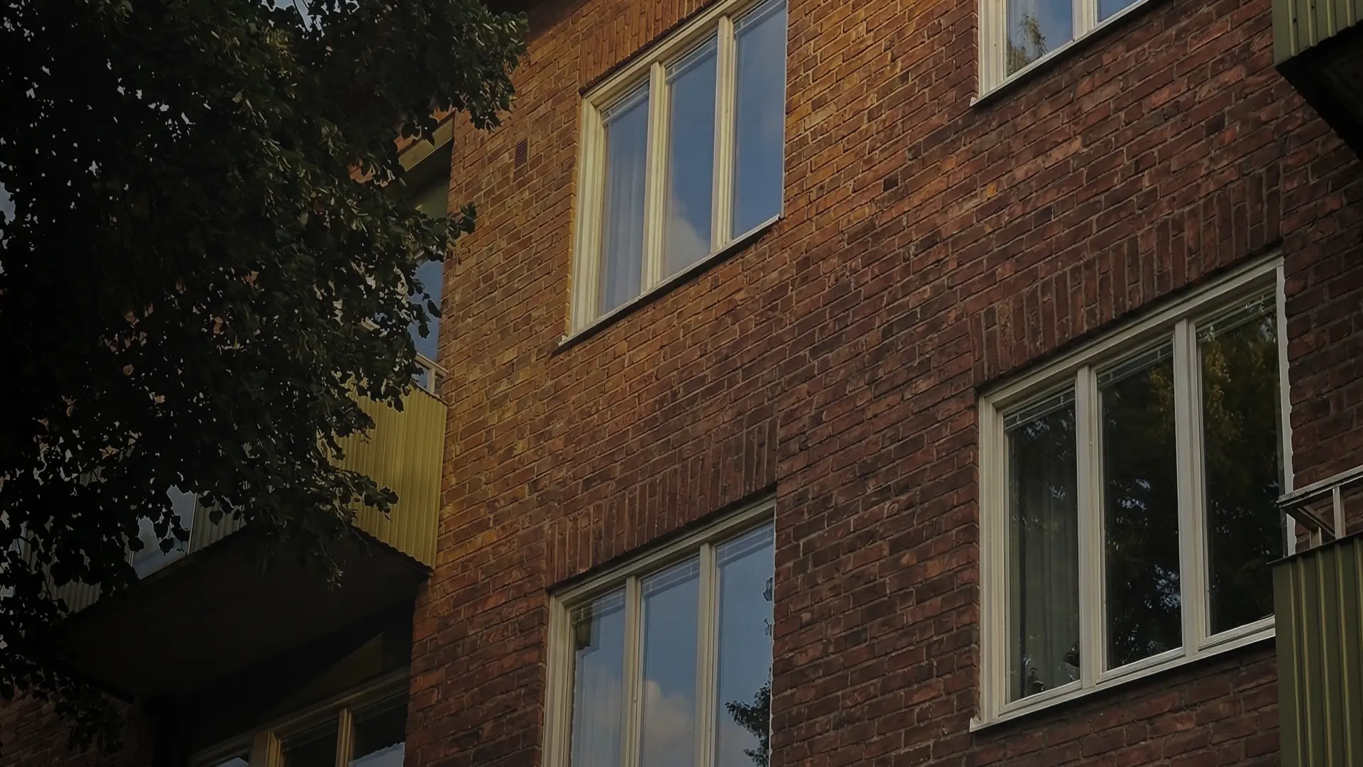 Fasad av flerbostadshus i rött tegel med flera fönster och balkong, omgiven av träd.