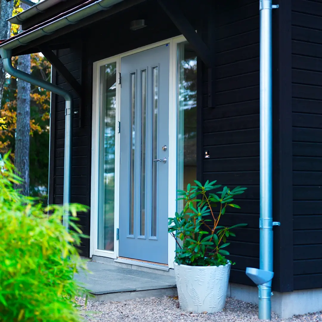 Grå ytterdörr med glaspartier på en mörk träfasad, med växt i kruka bredvid entrén.