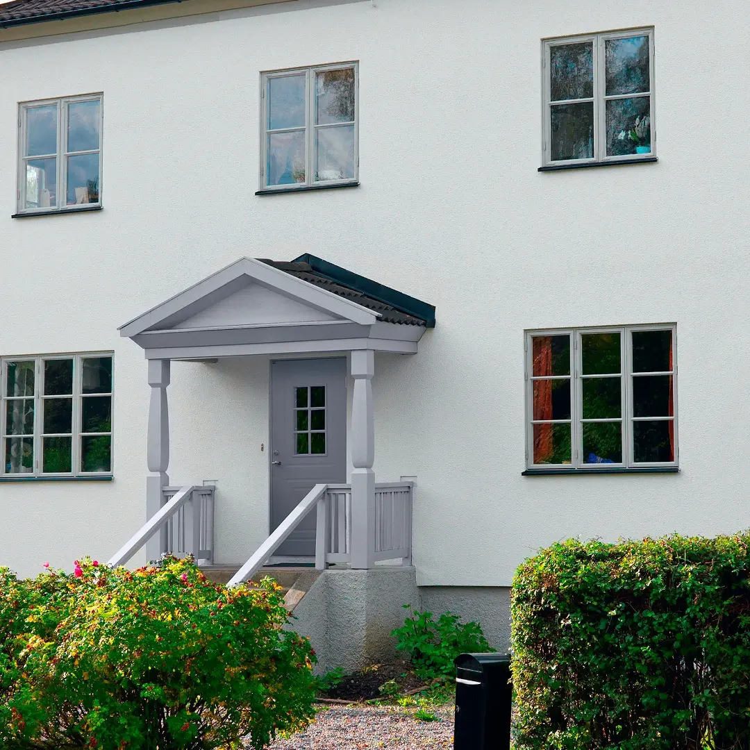 Framsidan av ett vitt tvåvåningshus med en liten täckt veranda, grå ytterdörr, symmetriska fönster och gröna buskar i trädgården.